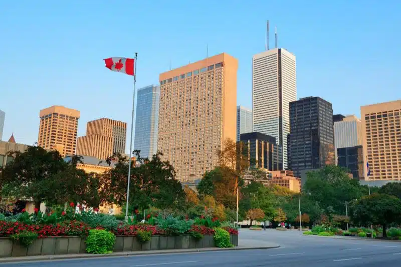 Toronto skyline with a prominent Canadian flag in the foreground, set against a clear blue sky and lush green landscape in the city center.