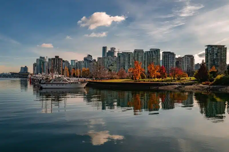 Vancouver skyline reflected on calm water with yachts docked in the marina and autumn-colored trees in the foreground under a partly cloudy sky