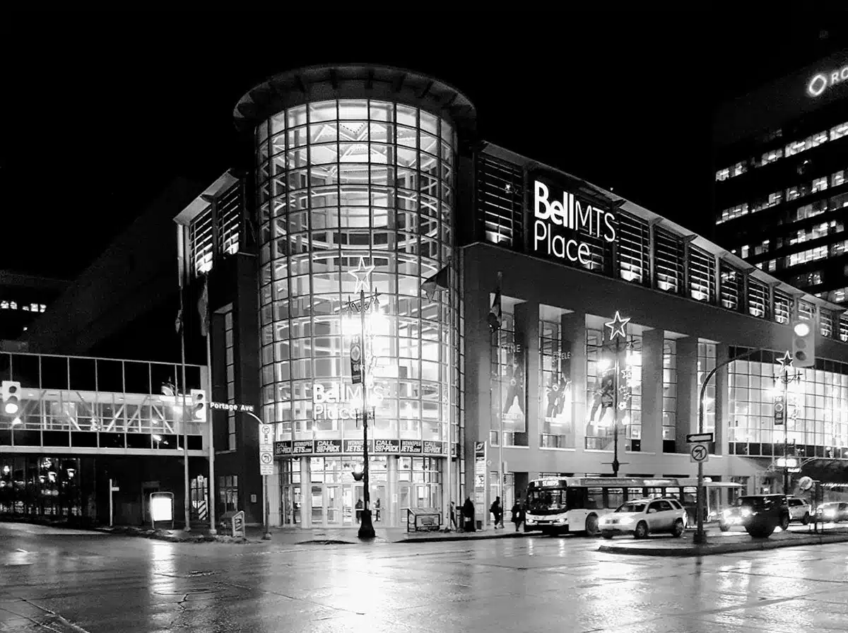 Black and white photo of Bell MTS Place at night, showcasing its illuminated glass entrance and surrounding city lights. The modern structure features a curved glass facade with "Bell MTS Place" signage above the entrance. Cars, a city bus, and pedestrians are visible in the foreground, adding to the urban nighttime scene.