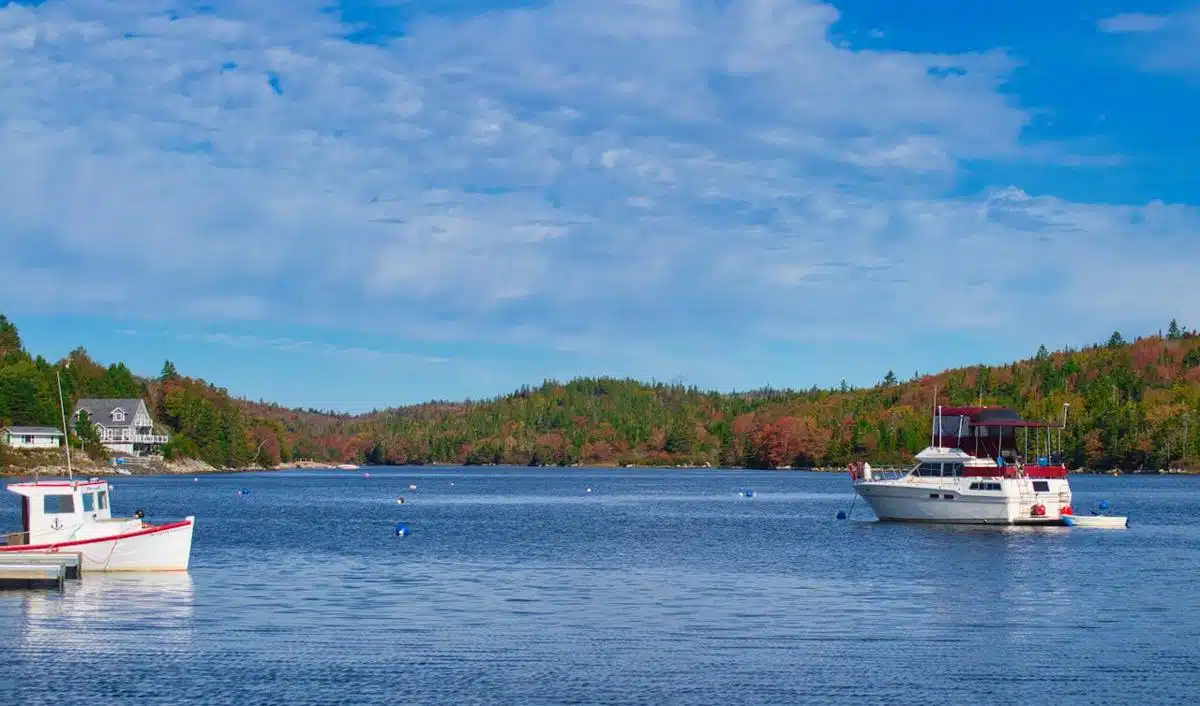 Scenic view of a tranquil lake near Halifax, Nova Scotia, with boats floating on the calm water. A small, white fishing boat is docked on the left, while a larger boat with a red canopy is anchored nearby. The background features lush, forested hills with autumn foliage under a bright, partly cloudy sky.