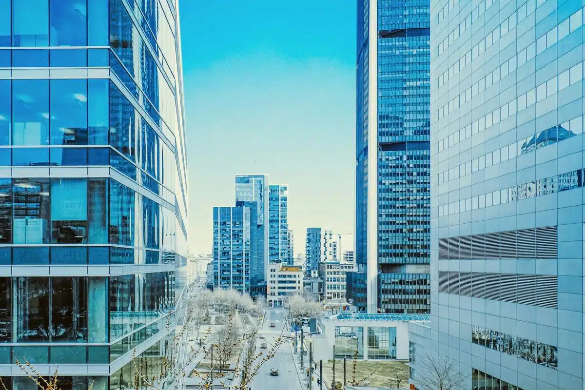Modern downtown Montreal cityscape with high-rise office buildings and glass facades, reflecting a clear blue sky. The street below is lined with trees, and the sleek architecture creates a vibrant, urban atmosphere on a bright day.