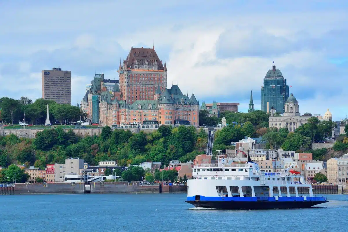 Iconic view of Quebec City with the Château Frontenac prominently standing above the waterfront, surrounded by historic buildings and lush greenery. A ferry crosses the St. Lawrence River in the foreground, with a partly cloudy sky framing the picturesque scene.