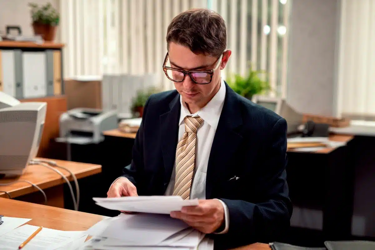 A tax lawyer, dressed in a suit and tie, carefully examines paperwork at his desk in an office setting filled with files and documents, reflecting a meticulous approach to financial review and legal documentation.