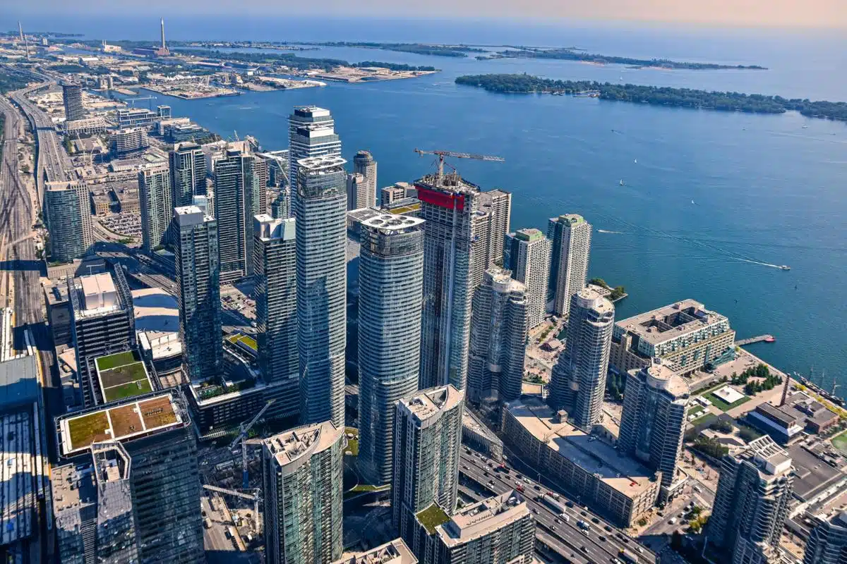 Aerial view of Toronto's downtown skyline featuring towering modern skyscrapers along the waterfront of Lake Ontario. The urban landscape is framed by the lake, with islands visible in the distance, creating a vibrant and expansive cityscape under a clear sky.