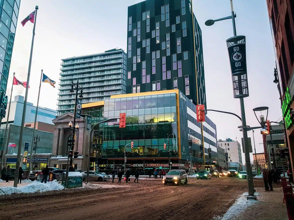 Busy downtown Winnipeg street scene with modern glass buildings and pedestrians crossing, framed by Canadian flags and street banners. The snowy roads and warm interior lights create a contrast, capturing the city's urban winter atmosphere.