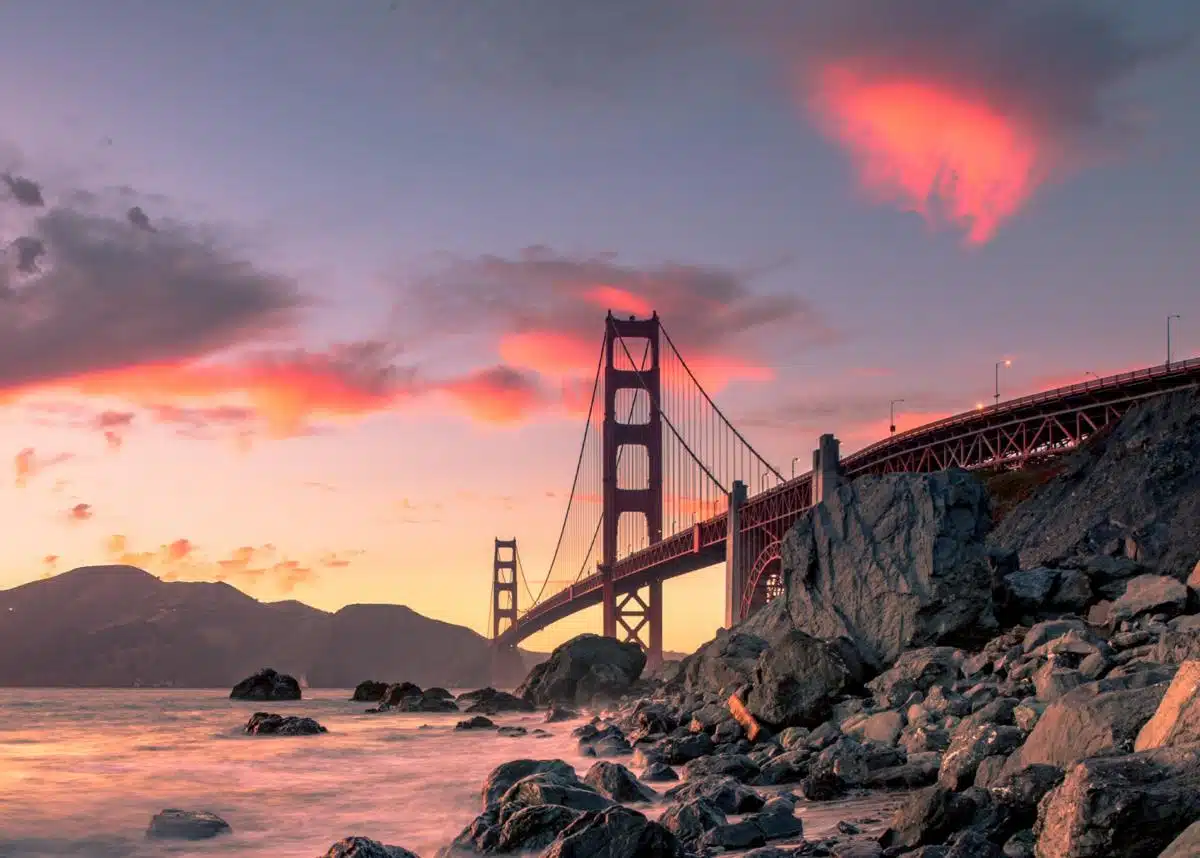 Scenic view of the Golden Gate Bridge in San Francisco, California, at sunset with rocky shoreline and colorful sky.