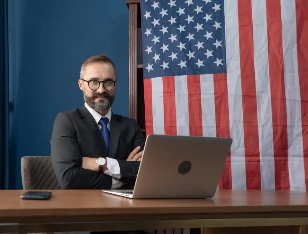 Confident tax attorney in Houston office with U.S. flag background and laptop on desk.