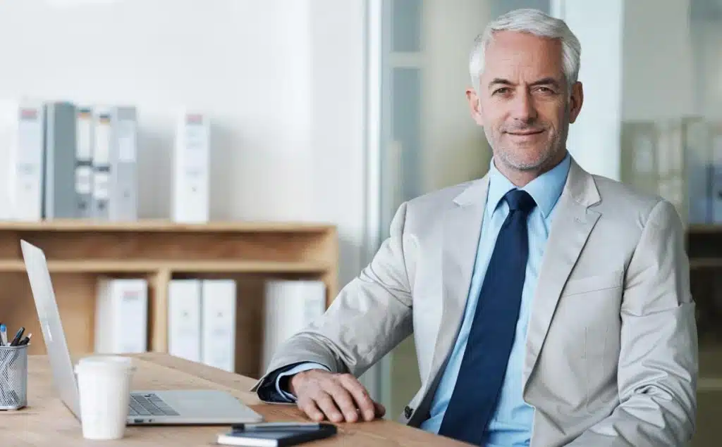 Confident mature tax attorney sitting at desk in Orlando office with laptop and coffee cup.