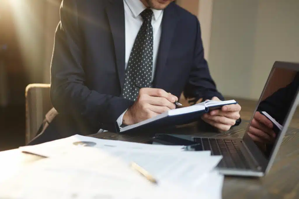 Close-up of a suited tax planning lawyer jotting notes in a blue notebook while reviewing documents beside an open laptop.