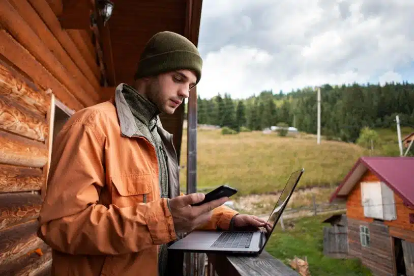 Man on a balcony in a rural setting using a laptop and a smartphone.