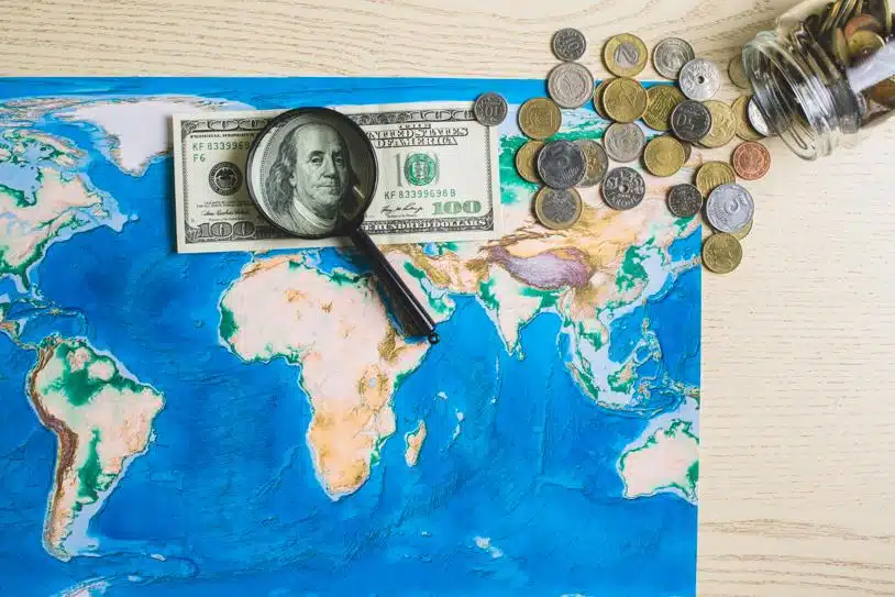 An American dollar under a magnifying glass on top of a world map beside coins from around the world spilling out of a jar.