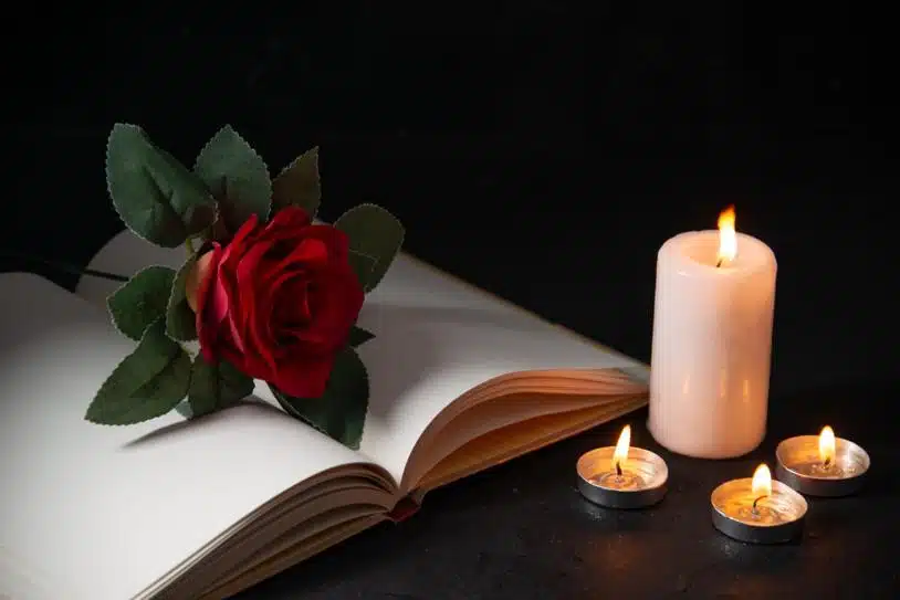An open, blank notebook with a rose placed on it beside candles against a black background.